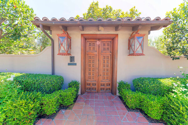Wooden door gate entrance with orange wall lamps and doorbell on the post at La Jolla, California. There is a red bricks entrance pathway in the middle of the shrubs at the front of the concrete wall.