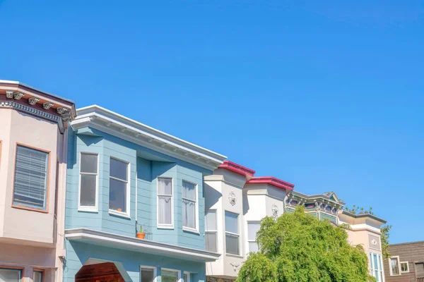 Row of houses with bow windows against the clear blue sky in San Francisco, California. Townhouses exterior with colorful palette and trimmings with a view of a tree at the front.