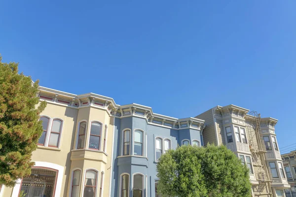 Complex townhouses with bow windows in a low angle view in San Francisco, California. There is a building on the right with emergency stairs and two buildings on the left with beige and blue exterior.