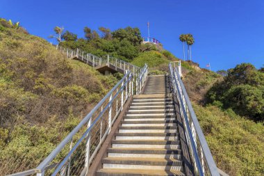 L-type outdoor staircase in San Clemente, California. Staircase with metal railings on a mountain slope with wild shrubs and trees against the clear sky.
