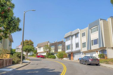 Curved road with double yellow lane in a residential area at San Francisco, California. There are cars parked on the side of the road near the suburban houses against the clear blue sky.