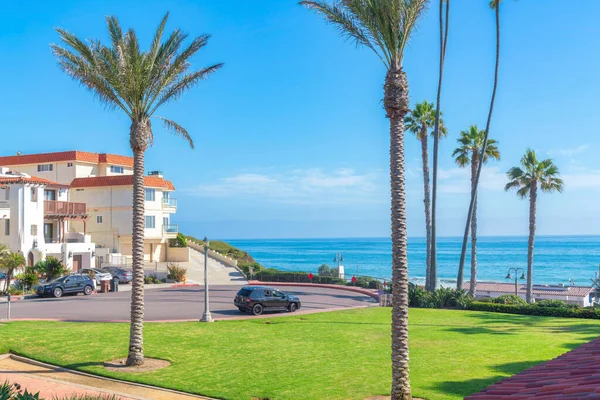 San Clemente community in California with a view of the blue ocean and clear sky. There are palm trees on the lawn at the front and vehicles on the concrete pavement near the buildings at the back.