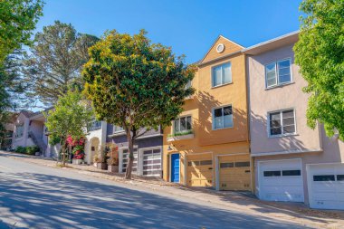 Sloped side walk and road with townhouses on the side in San Francisco, California. Exterior of residential buildings with garage and trees at the front along the side walk and driveways.
