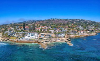 Aerial view of beachfront mansions in La Jolla, California. View of the blue ocean water at the front of the mansions and a view of beach houses against the clear blue sky at the back.