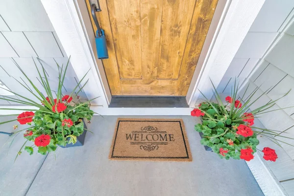 Welcome doormat in the middle of potted flowering plants near the wooden door with lockbox. Outside a house with a painted wood shingle siding and concrete entrance with brown doormat.