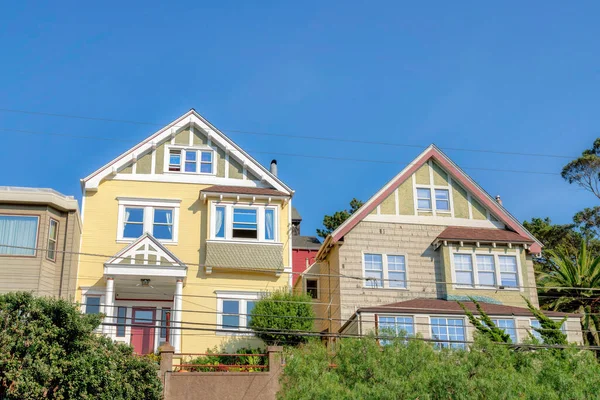 Two houses with gable roofs in the suburbs neighborhood in San Francisco, CA. There is a yellow house on the left with a view of red front door beside the house on the right with wood shingle sidings.