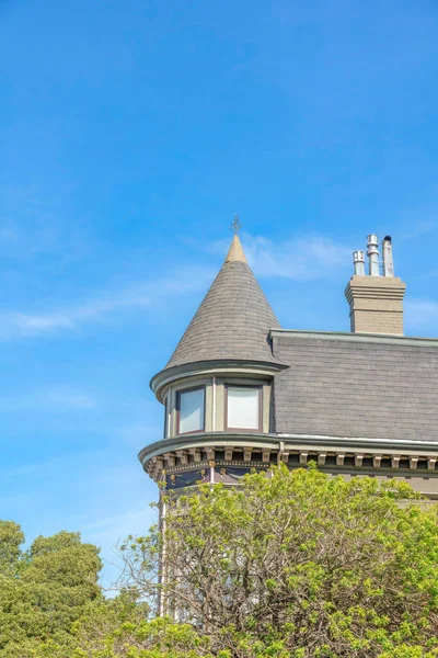 Trees below outside the french country home in San Francisco, California against the sky. Low angle view of a house with a view of the curved wall and gray shingles roof and chimney with flues.
