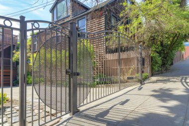 Wrought iron black gate of a residence in San Francisco, California. Front of a house with concrete driveway and a view of a tree near the house with wood shingles sidings at the back.