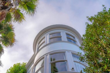 Low angle view of a curved wall of a house with reflective glass windows in San Francisco, CA. White residential building exterior with trees at the front against the sky.