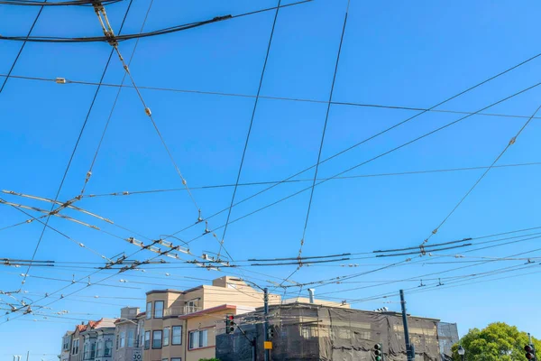 Cables above an intersection used to power the public cable cars of San Francisco in California. There is a row of buildings with unfinished building at the back near street lights and stop lights.