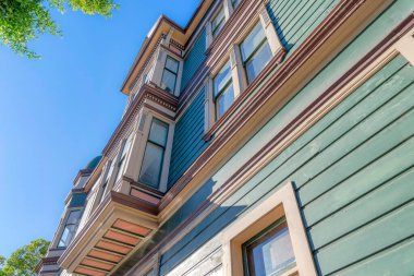 Low angle view of a victorian residential building with green wood lap sidings in San Francisco, CA. There is a view of a bow windows with beige window frames and brown trims.