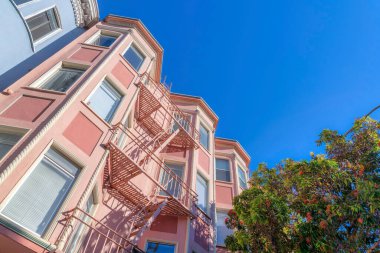 Low angle view of a pink apartment building with emergency staircase in the middle. Apartment building exterior with tall picture windows and tree outside on the right.