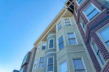 Three residential buildings in a row against the clear sky in San Francisco, CA. There is a building in the middle with beige shingles sidings beside the gray building and red violet wood shingles.