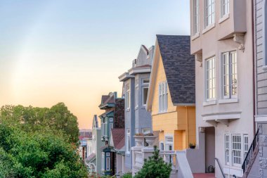 Row of houses and a view of the sunset sky in San Francisco, California. There is a house at the front with railings beside the yellow house with gray shingles sidings across the tree on the left.