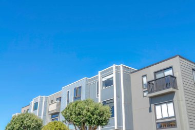 Townhomes with flat roof structures and gray wood siding in San Francisco, CA. There are trees at the front of the residential building with balconies against the clear blue sky.