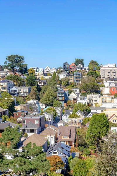 Single-family houses and apartment buildings on a slope in San Francisco, CA. Neighborhood with different residential architectural structures with trees in between against the clear sky background.