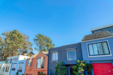 Row of small houses with reflective windows and attached garages in San Francisco, California. Facade of colorful houses against the trees and clear sky background.