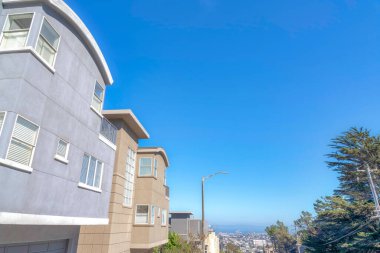 Three-storey houses with attached garages near a street light and electrical posts in San Francisco. There are houses on the left and trees on the right and a view of the urban area in the middle.