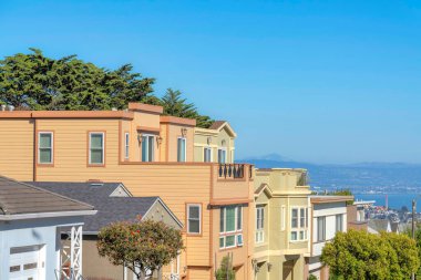 Three-storey suburban houses in a row with roof decks and a view of the bay in San Francisco, CA. Residential area on a higher area with row of single-family homes with attached garages.