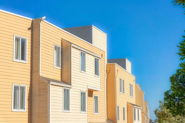 Apartment building exterior with yellow wood lap siding in San Francisco, California. Side view of an apartment building across the trees with casement windows against the clear sky background.