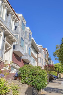 Sloped townhomes with planters and parked vehicle at the front in San Francisco, CA. Row of townhouses with wood siding and bricks against the clear blue sky background.