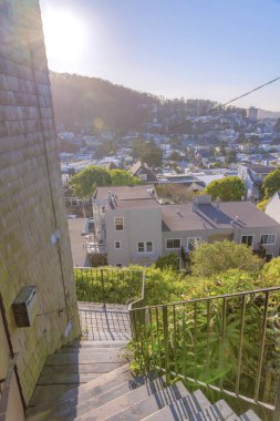 Sun peeking at the back of the building above the mountain in the neighborhood of San Francisco, CA. There is a stairs outside the building with wood sidings on the left and a view of houses below.