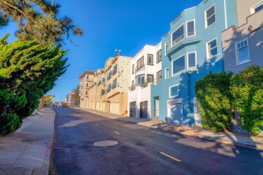 Street with yellow lanes at the front of large residential buildings in San Francisco, California. There are residential buildings on the left with attached garage on a sloped neighborhood.