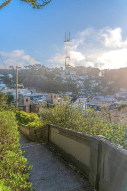 View of Sutro Tower against the clouds from a concrete pathway with barriers in San Francisco, CA. There is pathway at the front near the shrubs on the side with a view of the sloped residential area.