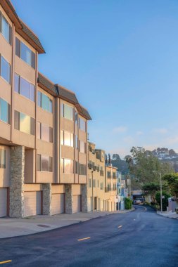 Row of apartment buildings with attached garages near the road in San Francisco, CA. There is an apartment at the front with reflective windows and a view of the trees and mountain at the back.