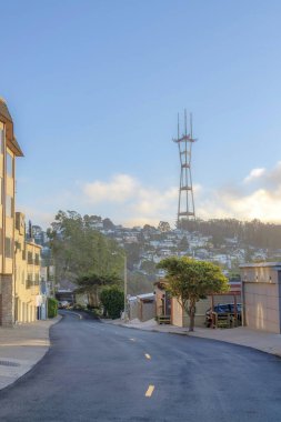 View of sloped residential area below the Sutro Tower in San Francisco, CA. There are houses on both sides of the empty road with trees and street lights.