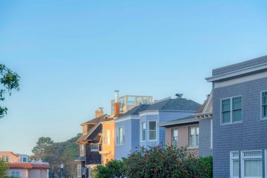 Row of houses with different exterior designs in the suburbs of San Francisco, CA. There are houses on both sides with wood shingle sidings and houses in the middle with painted concrete walls.
