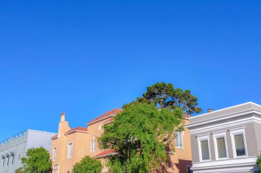 Three houses in a row at San Francisco, California against the sky. There is a peach house in the middle beside the gray houses with roof deck on the left and white trims on the right.