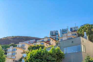 Apartment buildings on a slope of a mountain at San Francisco, California. There is a house on the left with wood siding and picture windows near the apartments with balconies and emergency stairs.