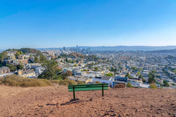 Lone green bench on top of a mountain with a view of buildings, and bay in San Francisco, CA. Rear view of a bench facing the cityscape against the mountains and skyline at the back.