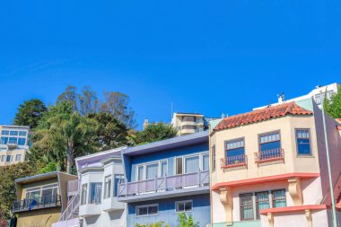 Facade of row of houses in San Francisco, California. There is a house on the right with railings on its picture windows and two purple houses in the middle near the box house on the left.