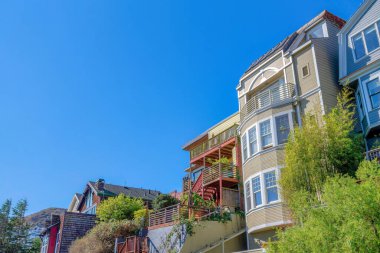 Low angle side view of multi-storey houses with wood sidings in San Francisco, California. There a house in the middle with stairs at the entrance and a view of a mountain slope at the left.