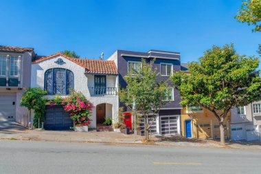 Sloped row of houses in San Francisco, California with trees and plants near the sidewalk. Facade of houses with attached garages near its front doors and painted exterior against the sky background.