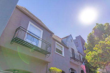 Sun glaring down directly at the houses in San Francisco, CA. There is a house on the left with window railings beside the house with USA flag below the window with railings and fake window shutters.
