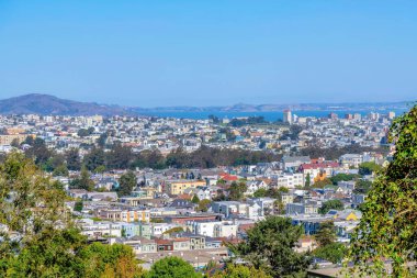 High angle view of San Francisco, California neighborhood near the bay. There are trees at the front of the view of the dense house buildings below and mountains against the sky at the back.