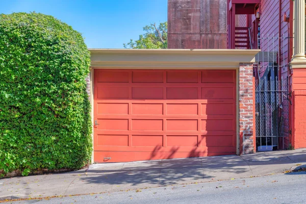 Detached garage exterior with peach wooden garage door at San Francisco, California. There is a shrub wall on the left of the garage and a gate on the right near the red building.