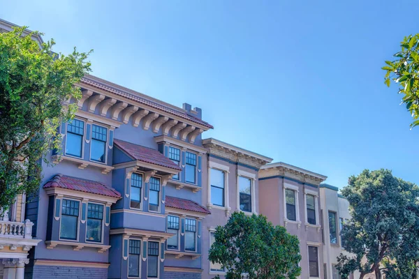 Adjacent victorian houses at San Francisco, California. There are trees at the front of the houses with decorative trims and reflective windows.