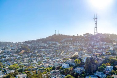 Dense housing on a sloped land in San Francisco, California below the Sutro Tower. High angle view of large residential buildings in a suburban area near the hills against the sun at the background.