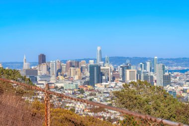 View from above of skyscraper buildings in the middle of midrise buildings in San Francisco, CA. There is a rusty wire fence and trees at the front of the cityscape near the bay at the back.