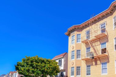 Aparment building with painted lined stucco wall and emergency ladder along with the suburbs houses. Neighborhood in San Francisco, CA with a tree at the front near the apartment building.