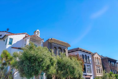 Trees at the front of mediterranean houses in a row at San Francisco, CA. There is a house on the right with wood shingle sidings along with the houses with stucco walls and balconies.