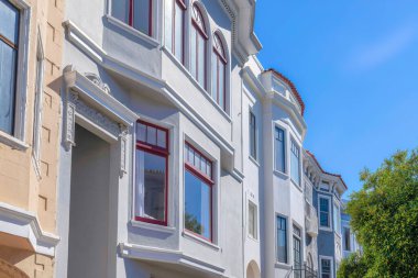 Rowhouses side view with painted walls across the trees against the sky in San Francisco, CA. Exterior of residential buildings with decorative trims and bay windows with relfective glass panes.