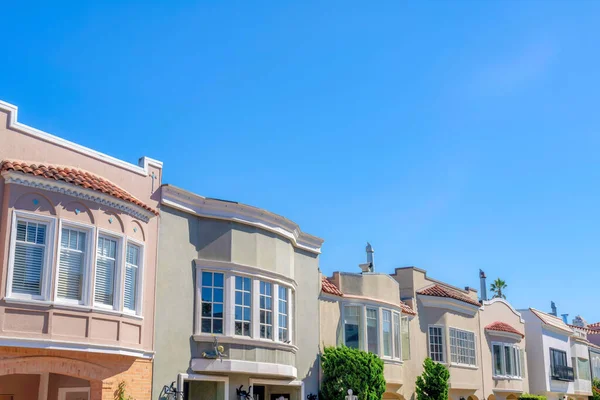 Row of painted houses with bow windows and clay tile roofs in San Francisco, CA. Exterior of houses with flues against the clear blue sky and a view of shrubs at the front.