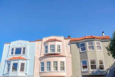 Three adjacent houses with clay roof tiles against the blue sky at San Francisco, California. There is a light blue house on the left beside the beige house near the gray house on the right.