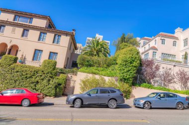 Dutch angle shot of a neighborhood with tilted houses in San Francisco, CA. There is a flat street at the front with parked vehicles against the large residential buildings with iron fence.