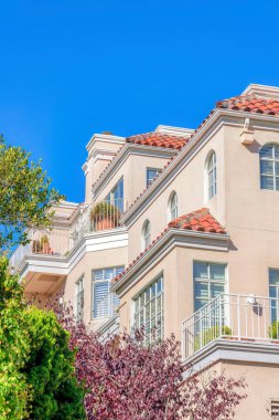 Side view of a modern mediterranean villa with potted plants on the balconies at San Francisco, CA. There are trees outside the house with paned windows and clay roof tiles.